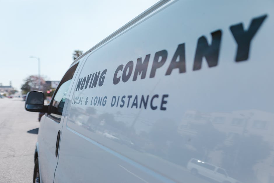 Image of three movers during a home relocation process, unloading large cardboard moving boxes from the back of a commercial moving van parked outdoors. The truck's open rear doors reveal stacking pla